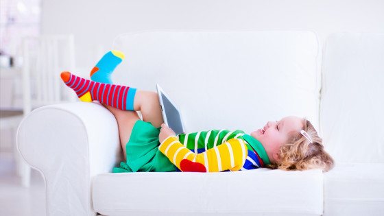 Little girl holding tablet pc relaxing on a white couch. Kids using computer at home or preschool. Children learning with digital devices. Child playing online game. Toddler kid and modern gadget.